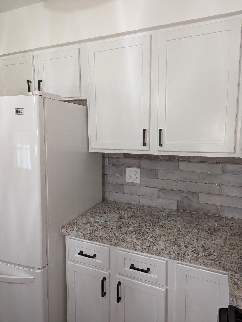 A white refrigerator stands next to kitchen cabinets with white doors, black handles, granite counters, and a tile backsplash.