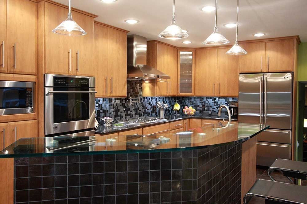 Modern kitchen with light wood cabinets, stainless steel appliances, a black tiled backsplash, and a glass-topped island.