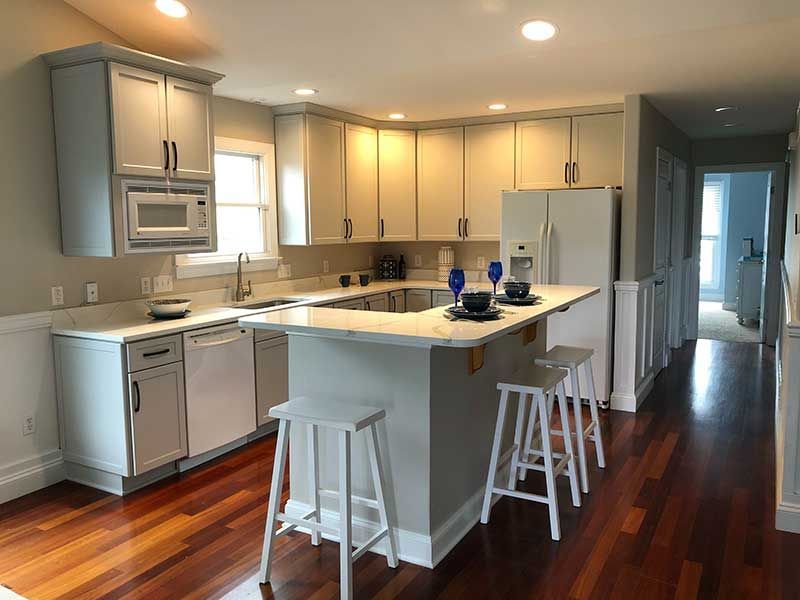 A modern kitchen with light gray cabinets, white countertops, a center island with two stools, and dark hardwood floors.