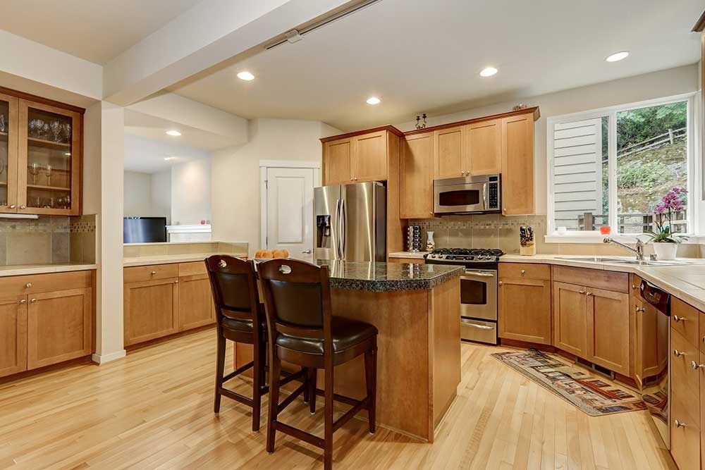 A kitchen with light wood cabinets, stainless steel appliances, a center island with two bar stools, and hardwood floors.