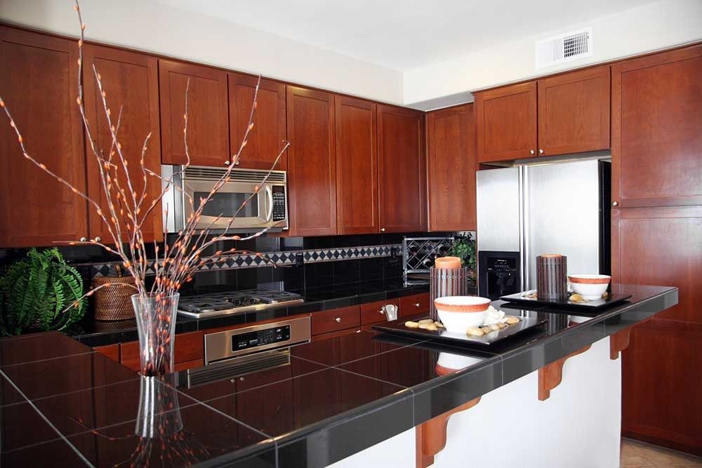 A kitchen with dark wood cabinets, black tile countertops, stainless steel appliances, and a decorative vase of twigs.