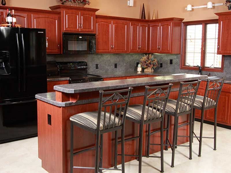 A kitchen featuring dark wood cabinets, black appliances, a raised breakfast bar with four stools, and a gray backsplash.