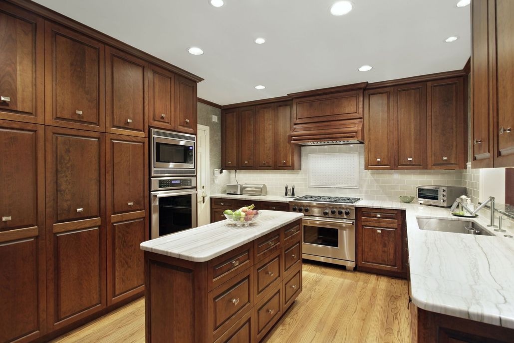 A warm-toned kitchen with dark wooden cabinets, white marble countertops, a central island, and stainless appliances.