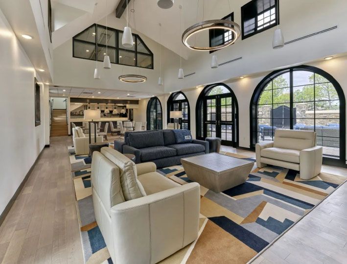 A bright, modern lounge area with high ceilings, large arched windows, and a patterned rug with couches and chairs.