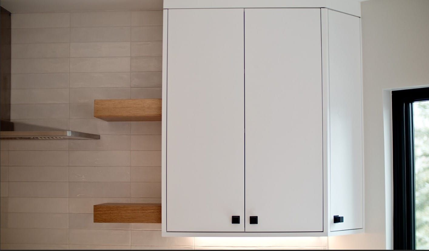 White kitchen cabinet with black square knobs, above two wooden shelves, against beige tile.