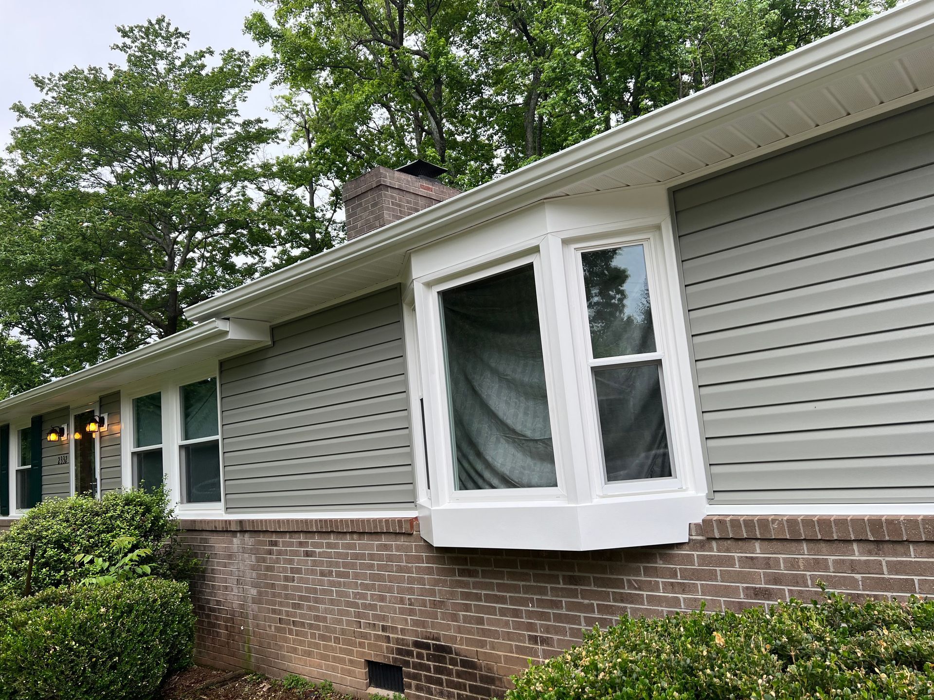 A gray-sided house with a white bay window and a brick foundation exterior, surrounded by trees and green bushes.