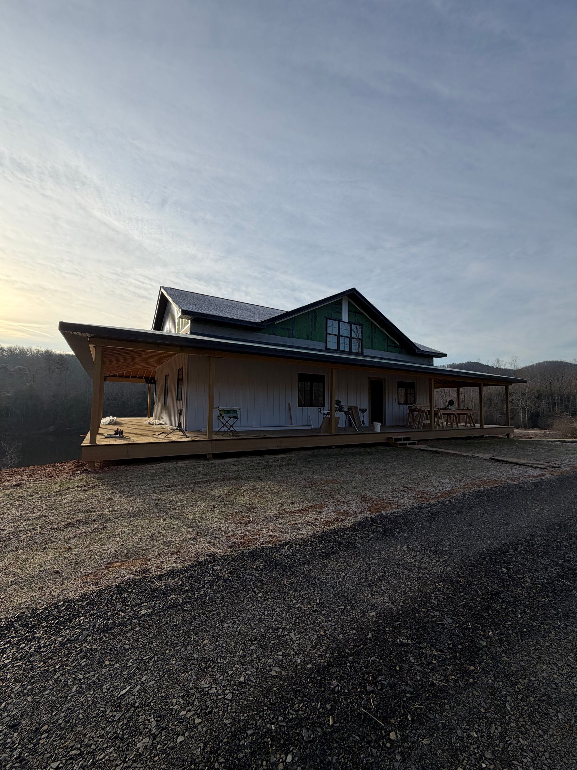 A single-story house with a wide porch and green trim sits on a gravel lot under a clear, pale sky at sunset.