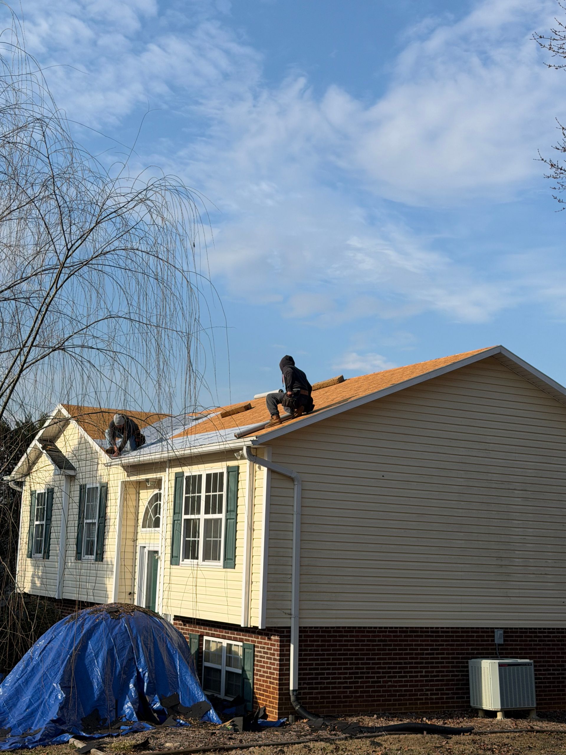 Two workers in black clothing install orange shingles on the roof of a beige house with a blue tarp on the ground.