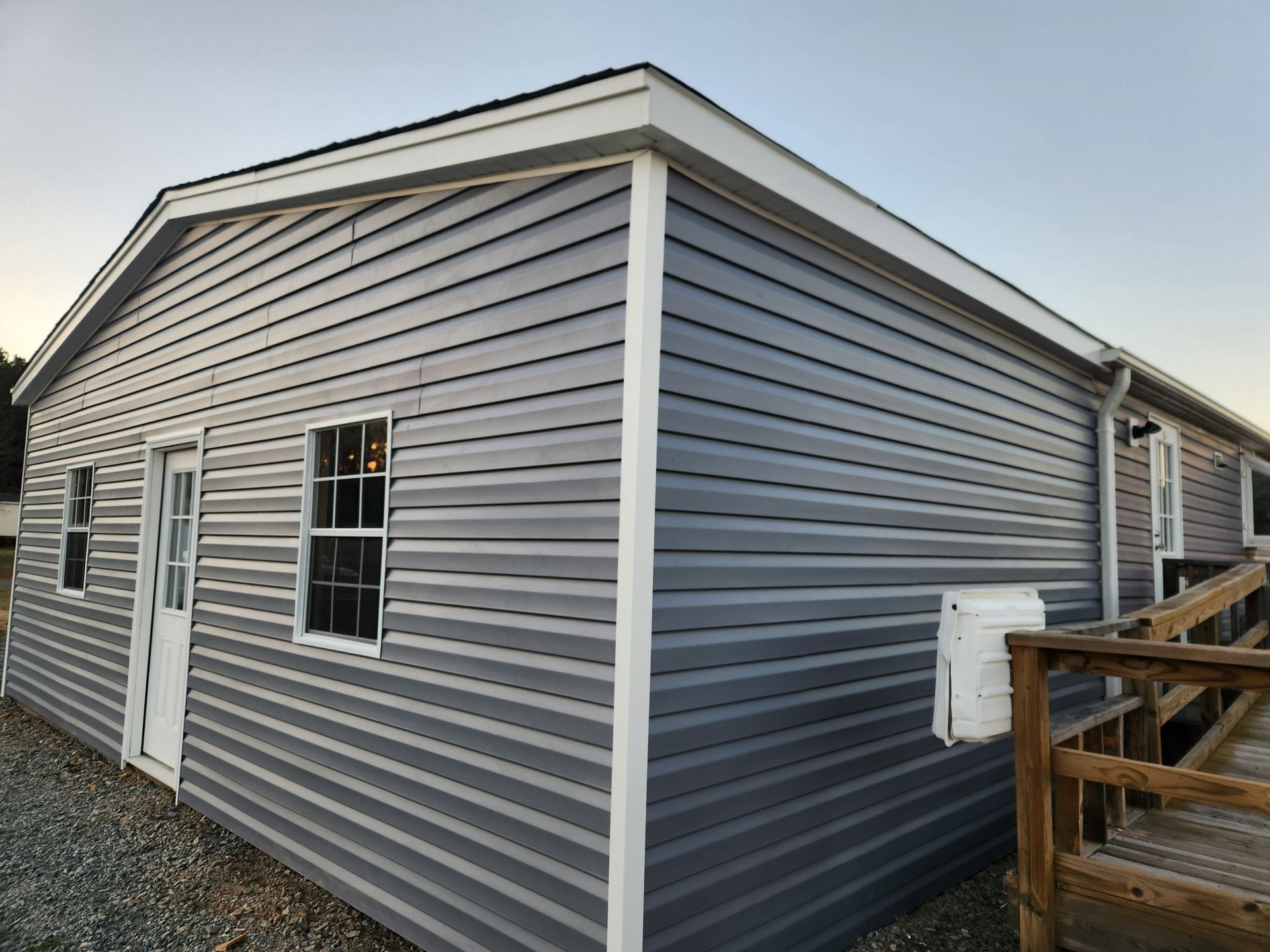 A gray, vinyl-sided modular building with a door, white trim, and an attached wooden ramp on a gravel surface.