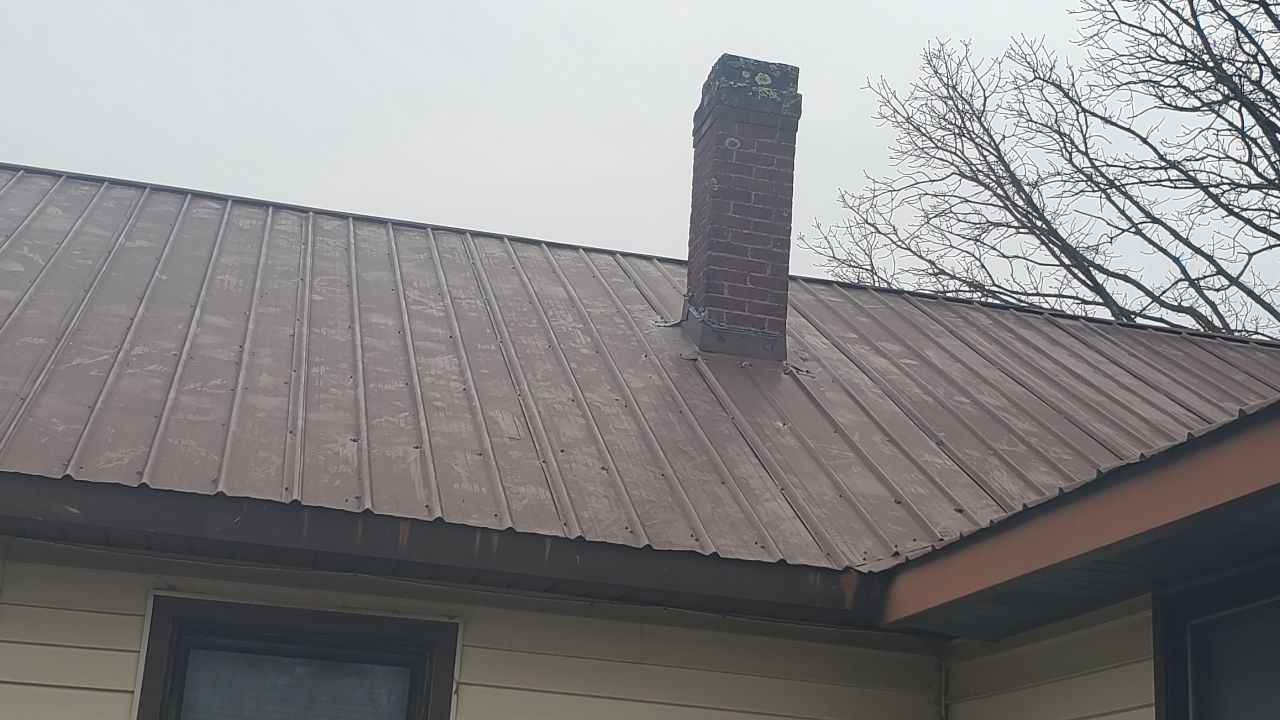 A brown metal roof with a weathered brick chimney extending upward against a cloudy sky.