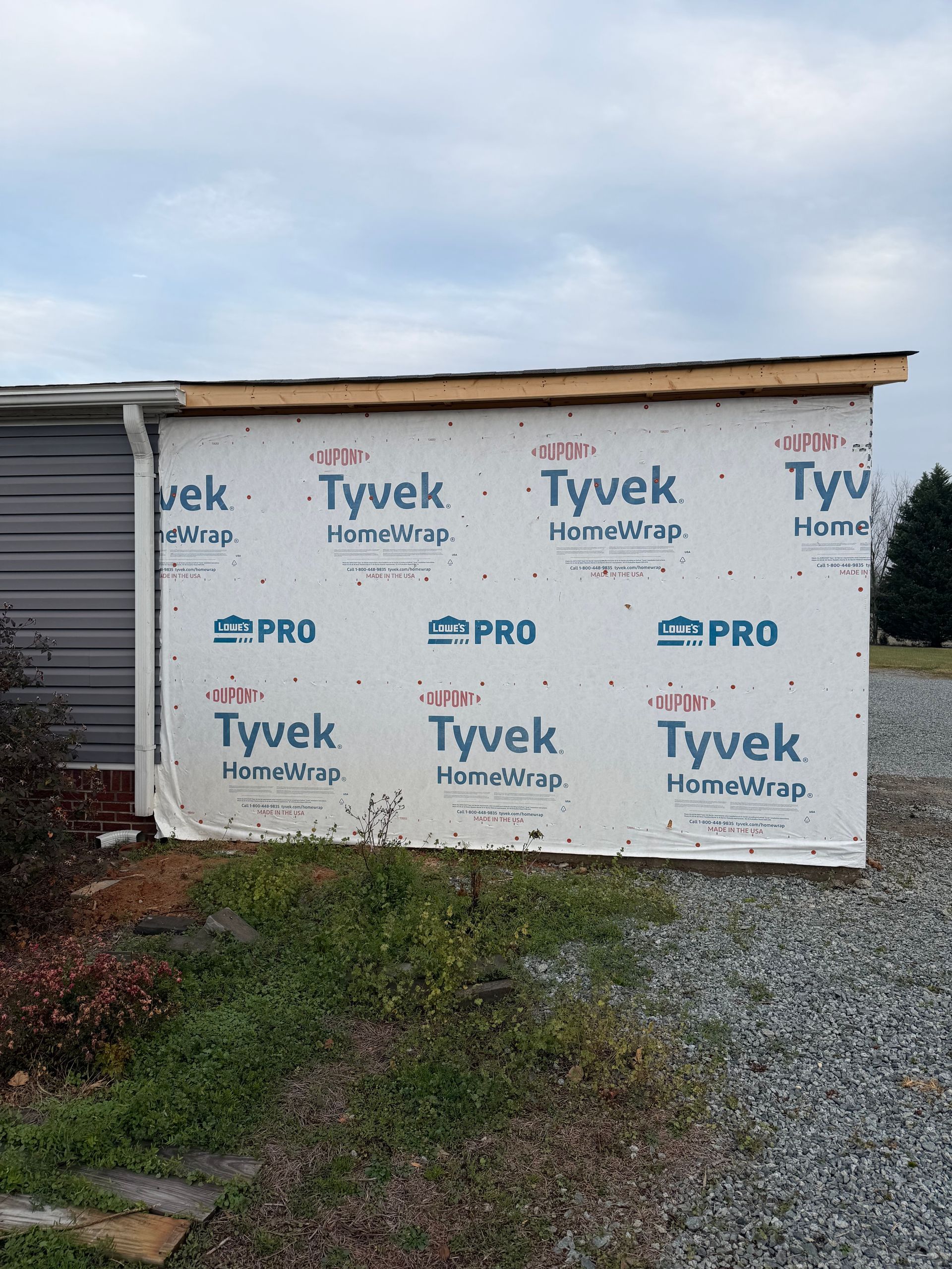 Exterior wall of a building undergoing renovation, covered with white Tyvek HomeWrap and located outdoors on a gravel lot.