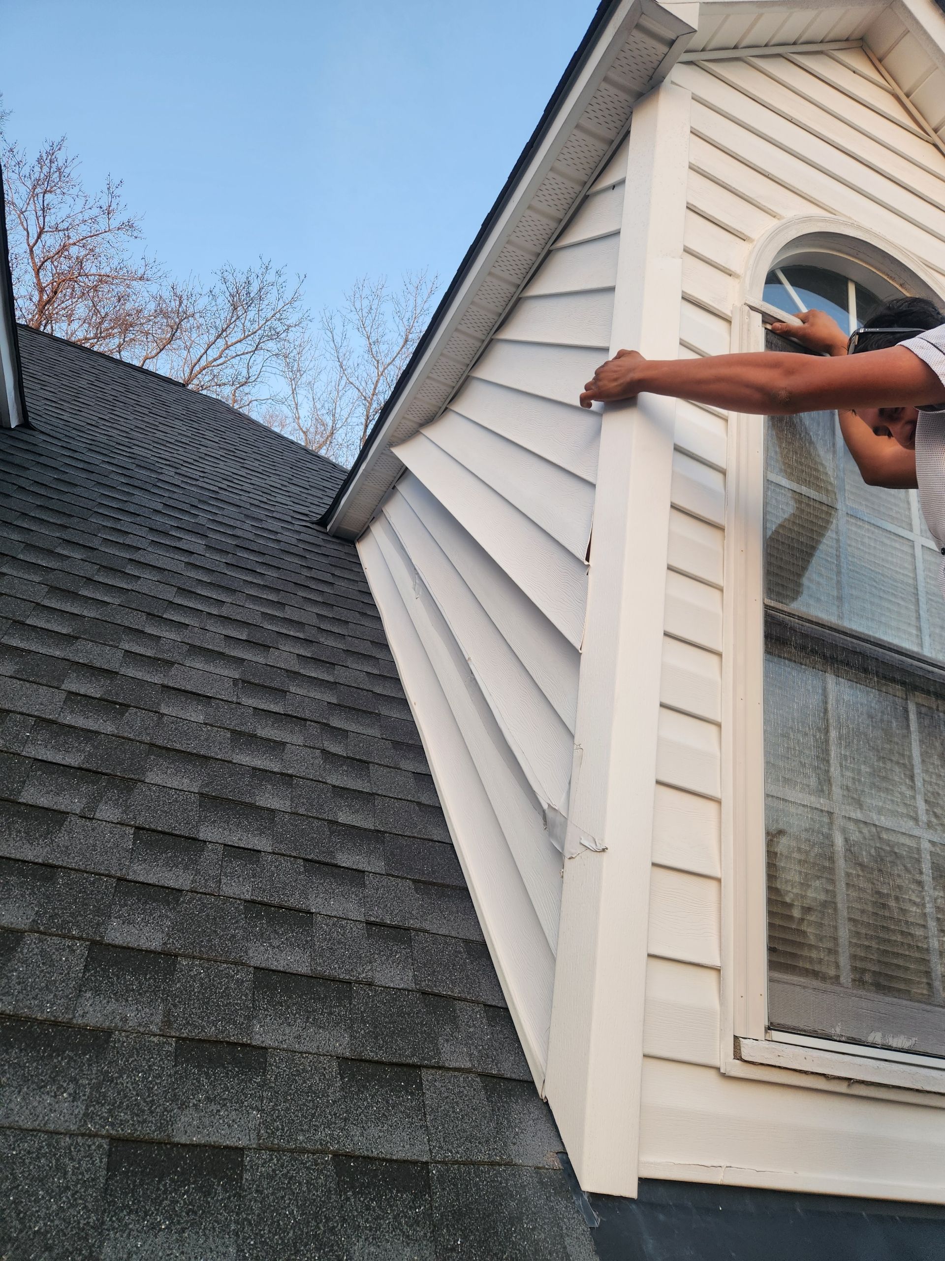 A person’s arm and hand use a tape measure to measure the corner trim of a white-sided dormer window on a shingled roof.