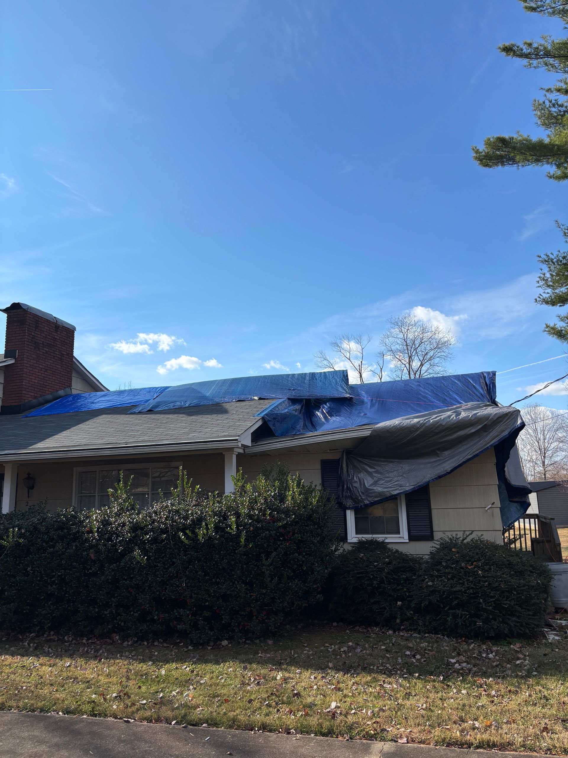 A house with a damaged roof partially covered by a large blue tarp under a clear blue sky.