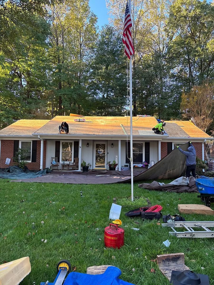 Workers are removing old roofing shingles from a house, with construction materials and an American flag in the yard.