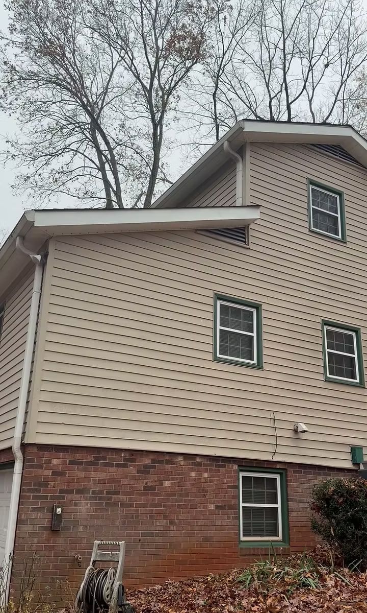 A side view of a two-story house with beige siding on the upper levels, a brick lower level, and windows, against trees.