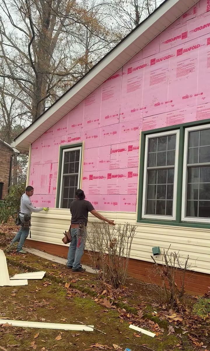 Two workers install cream-colored vinyl siding over pink insulation board on the side of a house with two windows.