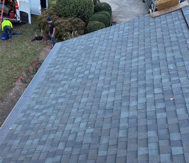 Workers install gray asphalt shingles on a residential roof, with a van and landscaping visible in the background.