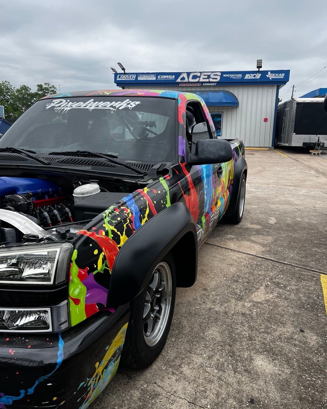 Truck with paint splatters, parked in front of a building. The hood is open, revealing the engine.