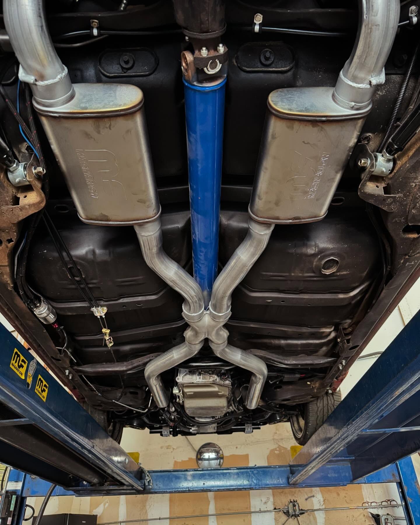 Undercarriage of a vehicle with custom exhaust system. Silver mufflers, blue driveshaft, black chassis.