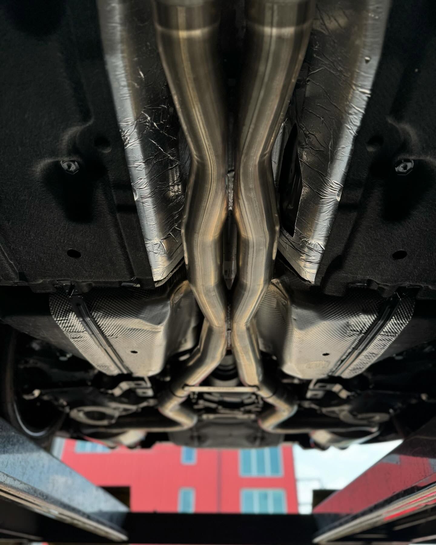 Undercarriage view of a car, showing a custom exhaust system. Silver pipes converge in an X-shape. The car is on a lift.