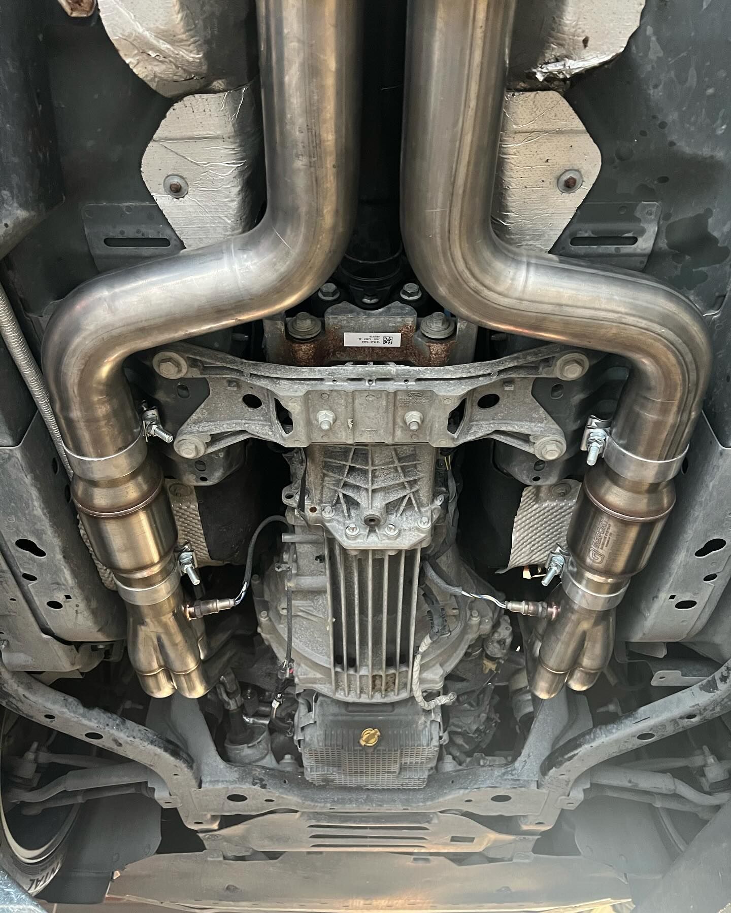 Undercarriage view of a vehicle showing the exhaust system and drivetrain components in a gray and silver setting.