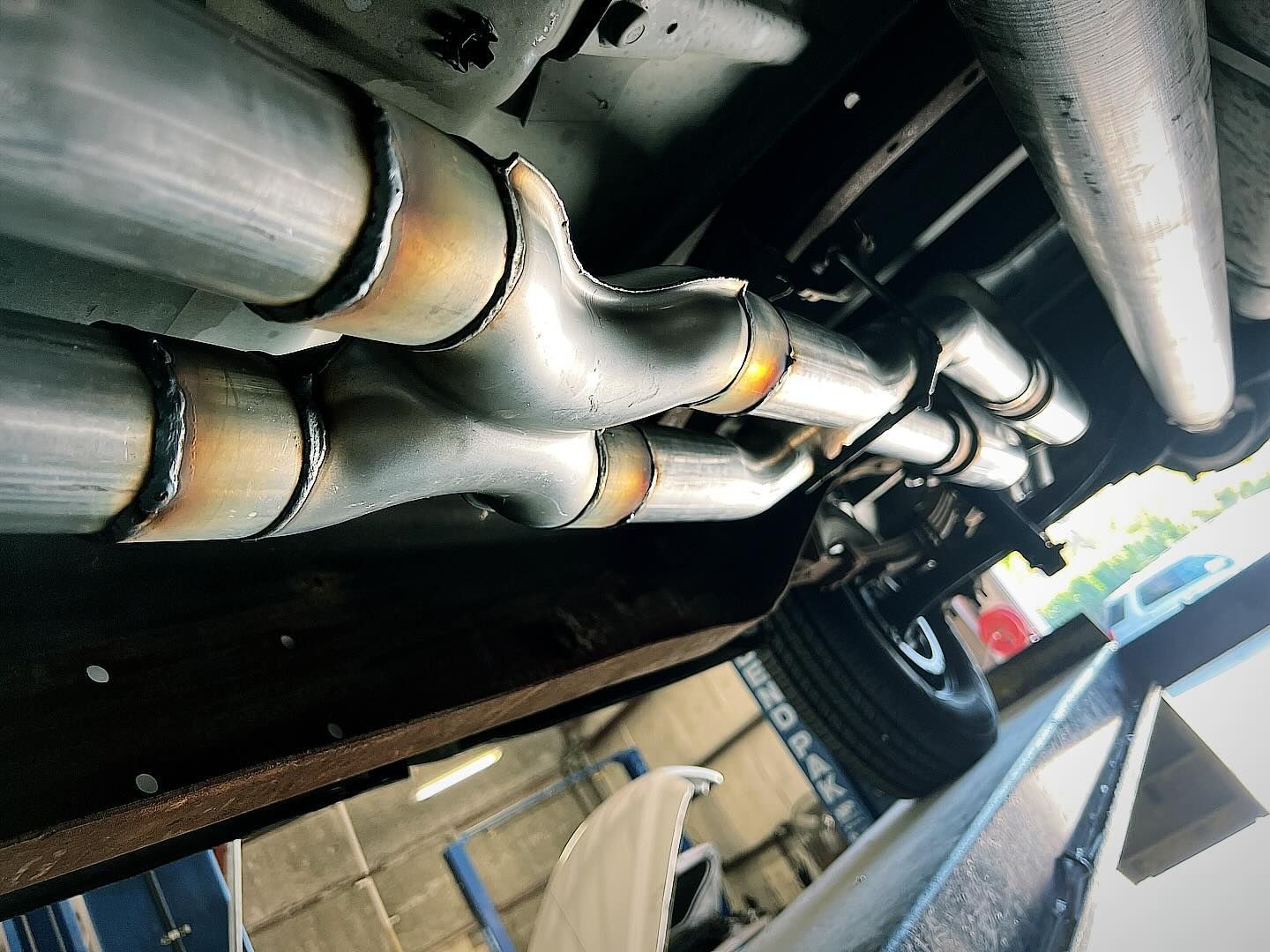 Undercarriage view of a vehicle's exhaust system with welded pipes forming an X-pipe configuration, visible from below.