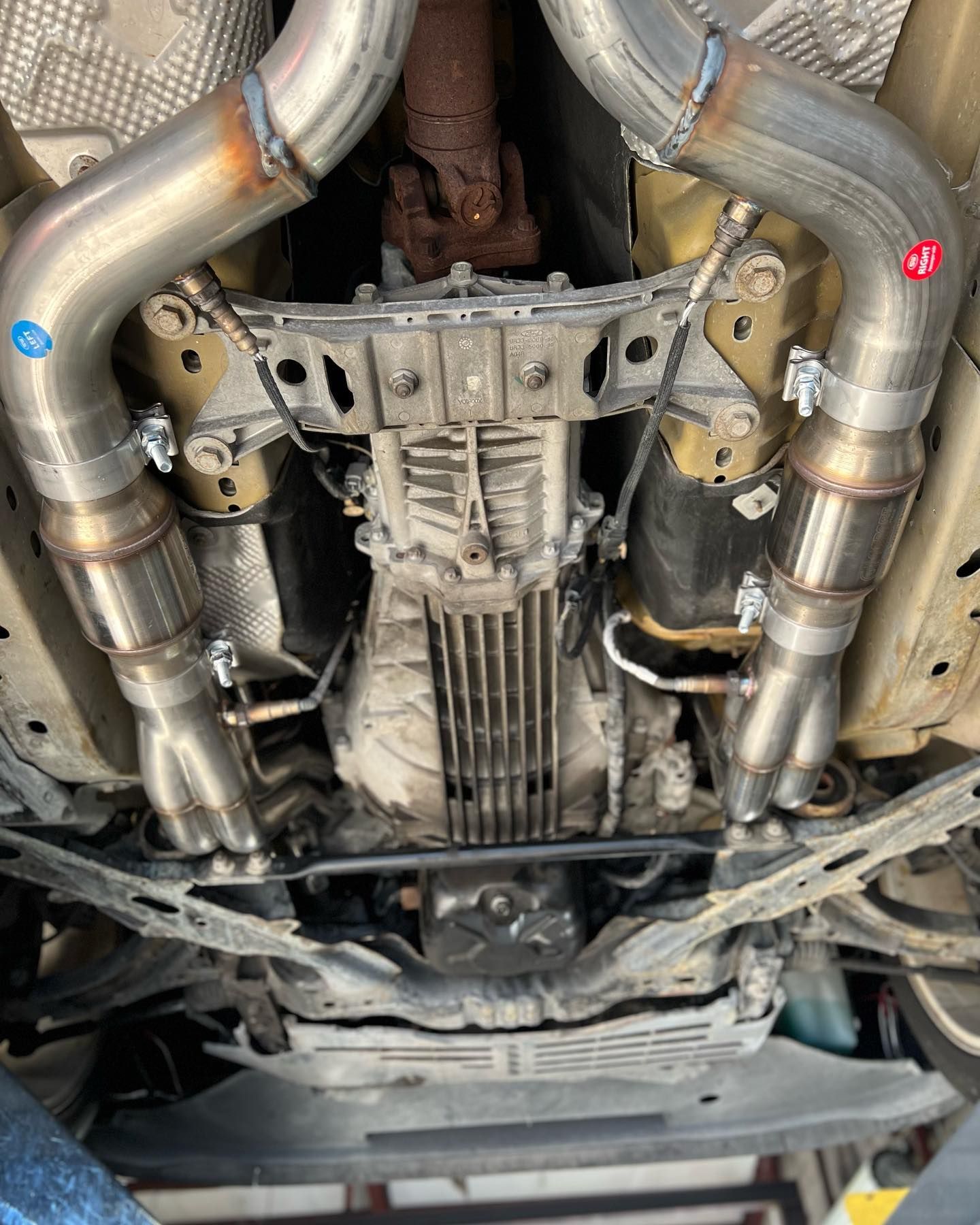 Underside of a car showing a custom exhaust system installation with metal pipes and clamps, view from below.
