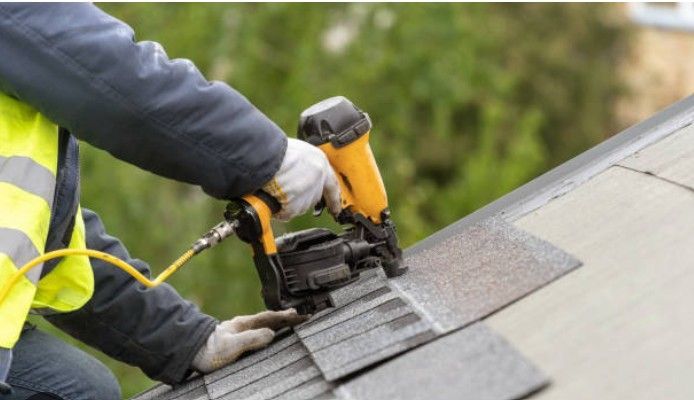 A construction worker in a high-visibility vest uses a yellow pneumatic nail gun to install shingles on a roof.