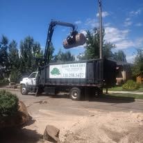 Two men stand near a truck and house; one wears a blue shirt and hard hat, the other a dark shirt and sunglasses.