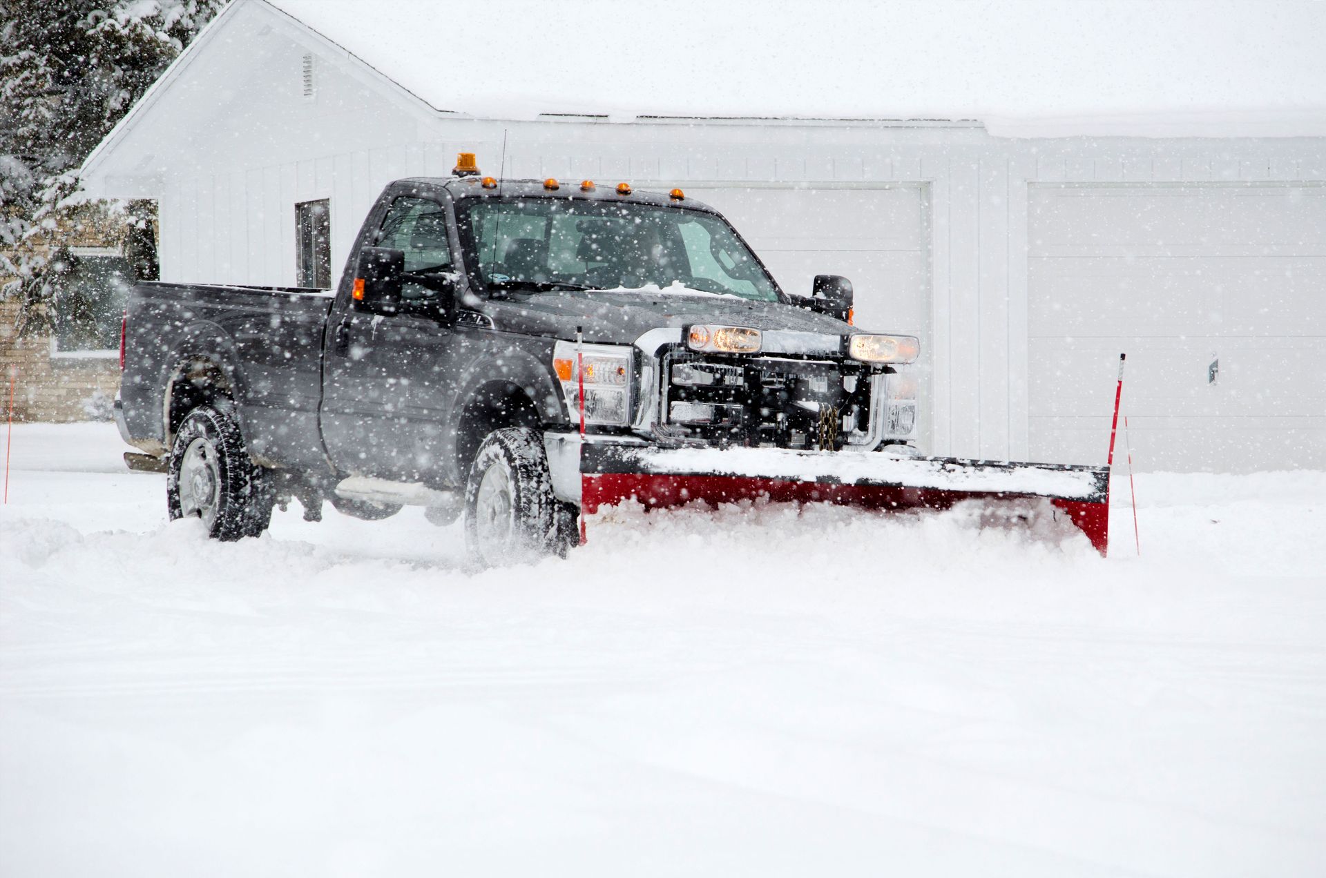 Black pickup truck plowing snow in front of a white house during a snowfall.