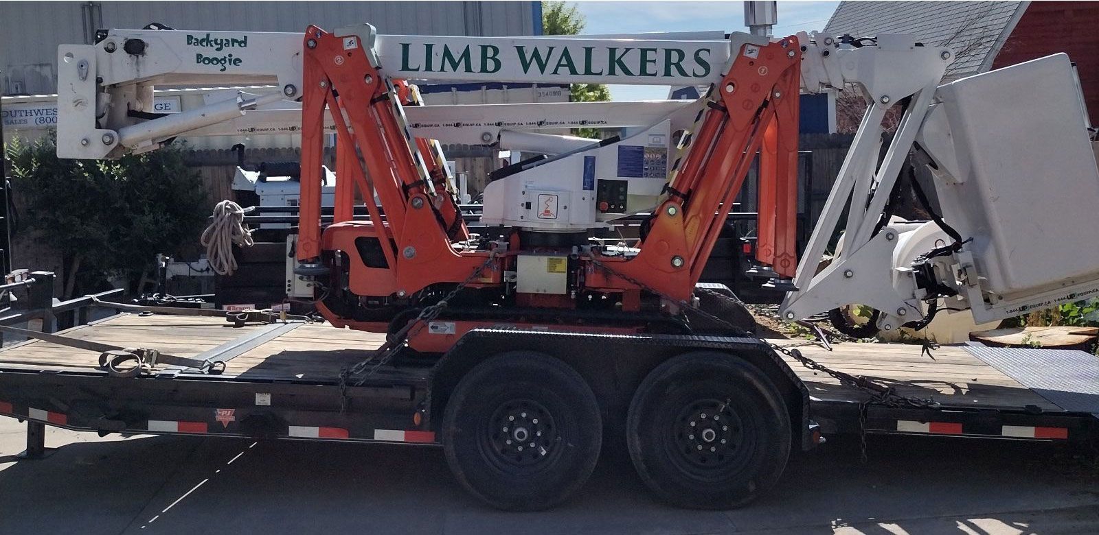 Orange and white tree trimming lift on a trailer, parked in front of a building. 