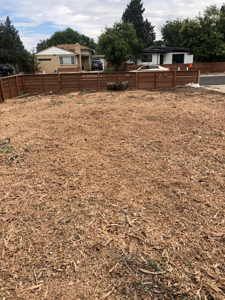 Wood-chipped lot behind a brown fence, with houses and a road in the background under a cloudy sky.