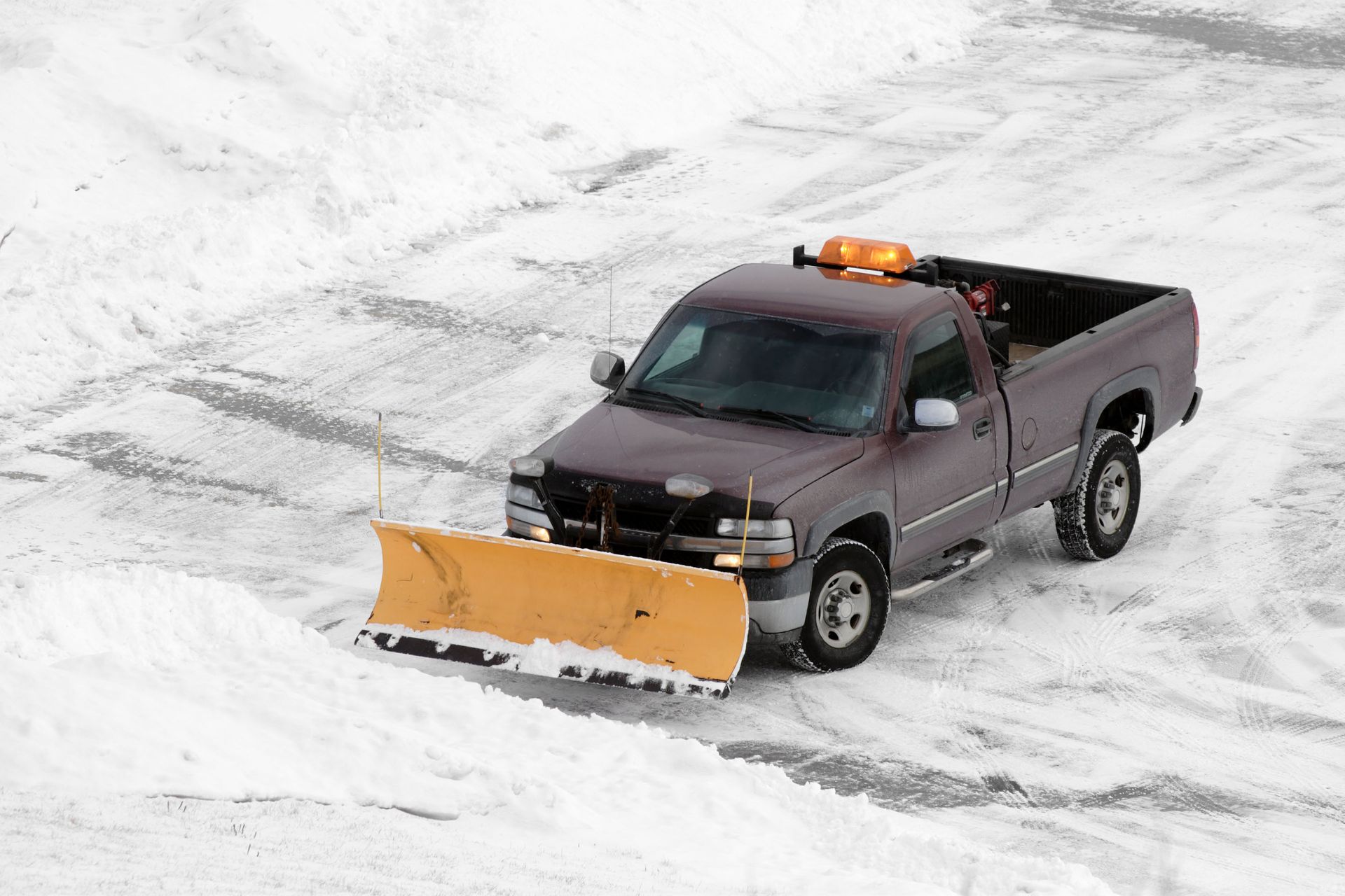 A maroon pickup truck with a yellow snowplow clears snow from a parking lot.