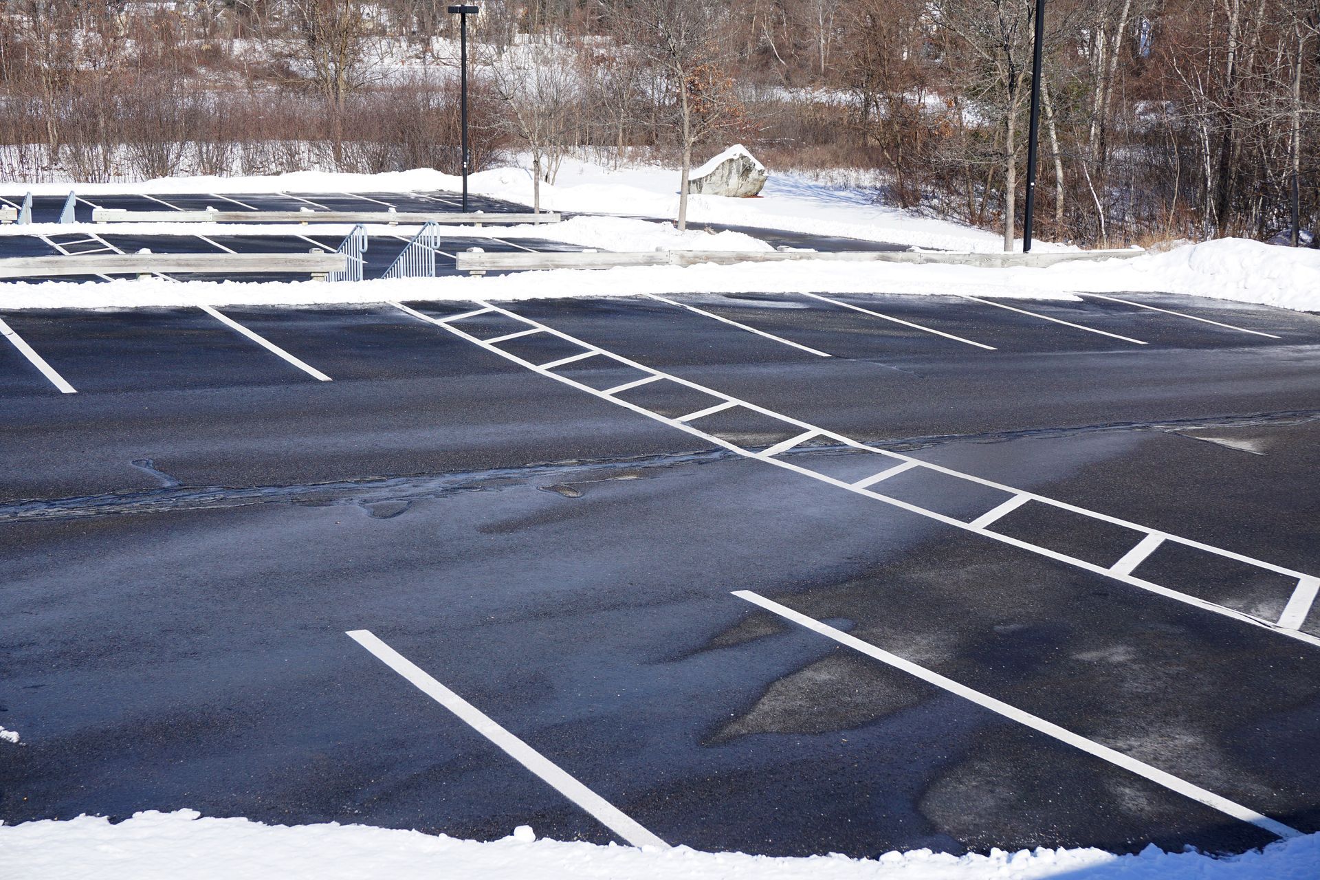 Snowy parking lot with painted white lines, partially covered in snow, leading towards a wooded area.