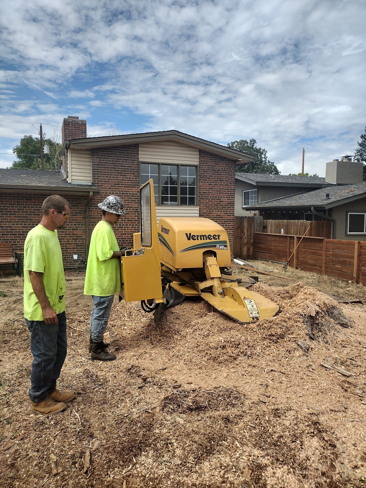 Two workers operating a yellow stump grinder, covered in wood chips, near a house.