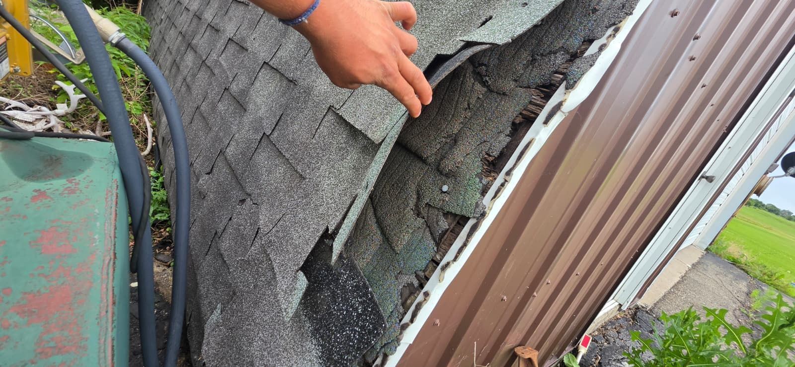 Hand inspecting damaged roof shingles beside a corrugated metal wall near a gutter