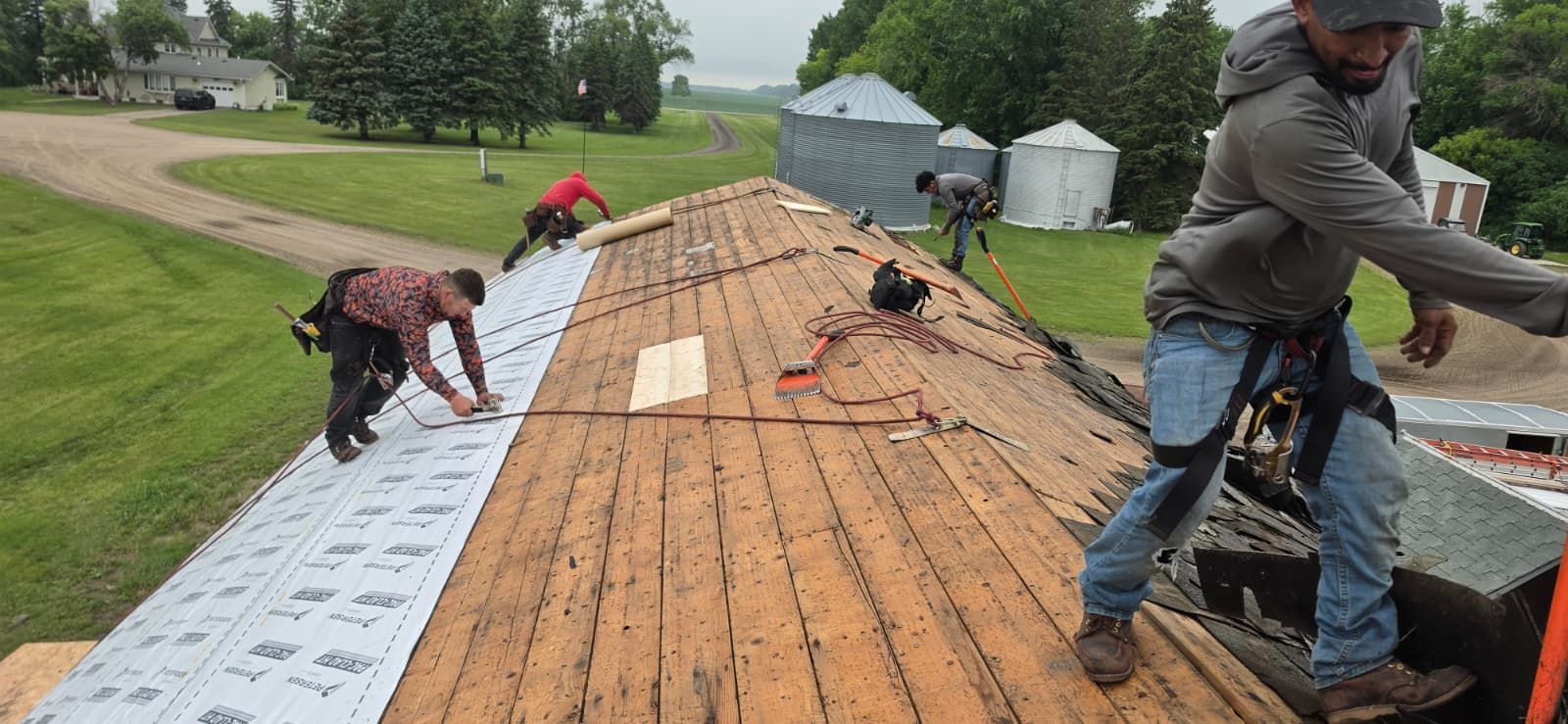 Workers installing shingles on a roof, with a ladder and open field in the background