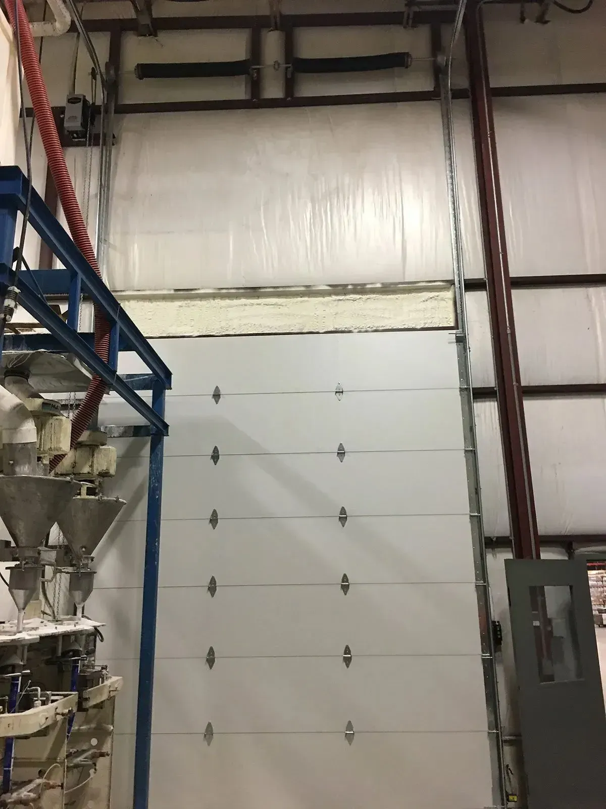 White overhead garage door inside a warehouse. Steel supports, equipment, and a small door are visible.