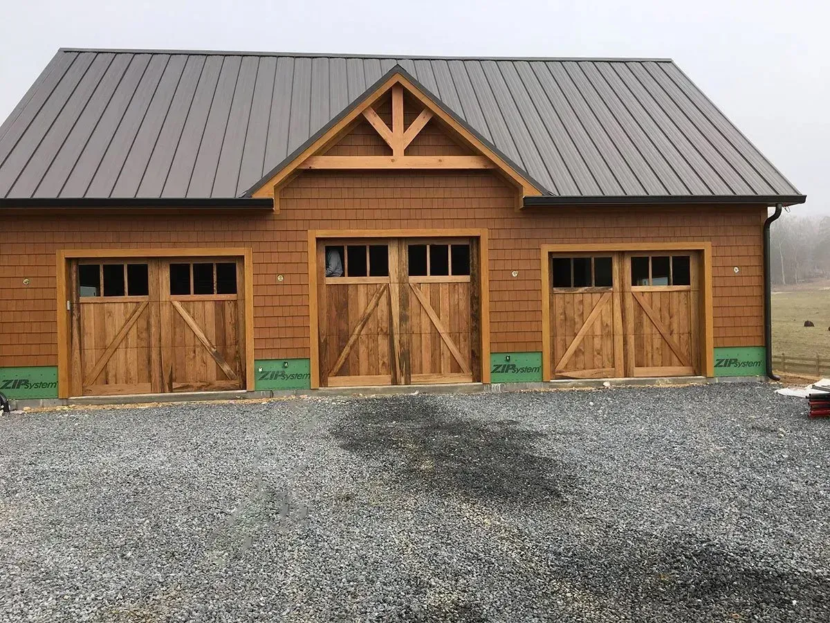 Three-bay wooden garage with rustic doors, gray metal roof, and gravel driveway.