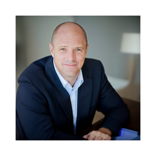 Man with a bald head in a blue blazer and white shirt, smiling, indoors with lamp in background.
