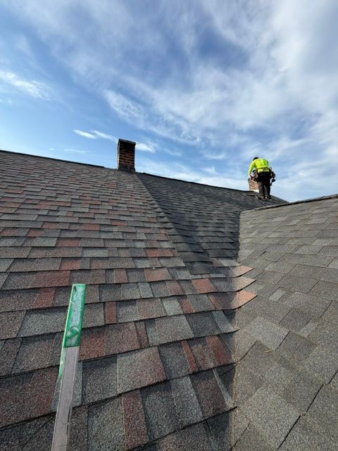 Worker inspecting a shingled roof under a cloudy sky, with a chimney and ladder visible