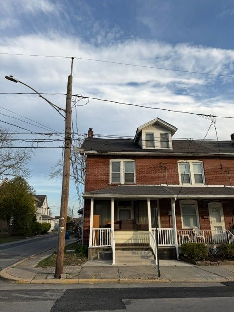 A red brick corner house with a front porch and dormer, beside utility poles on a cloudy street.