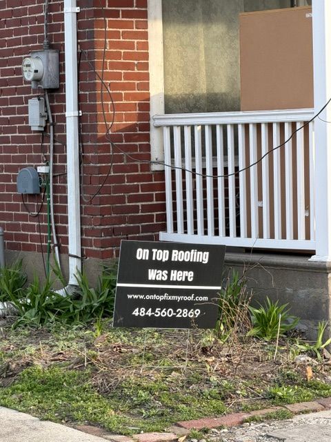 Roofing company sign beside a brick building and white railing, with weeds in the foreground.