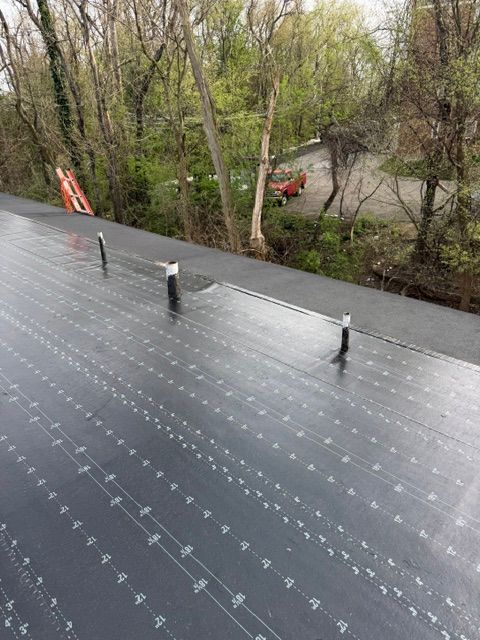 Black roofing membrane with three vent pipes on a roof edge, overlooking trees and a road below