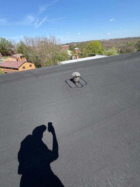 Flat black rooftop with a vent, overlooking trees and houses under a blue sky; shadow of person taking photo