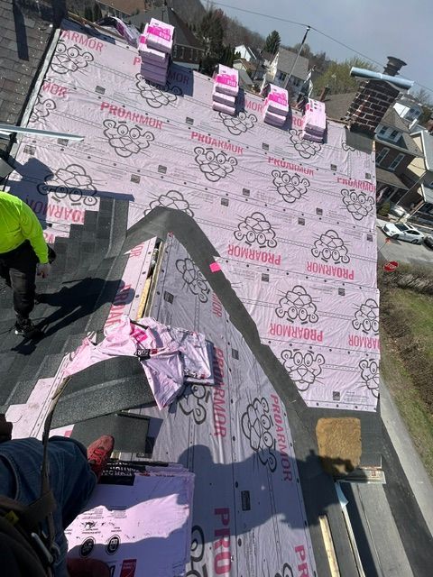 Roofer installing pink underlayment on a steep roof, with tools and a worker visible.