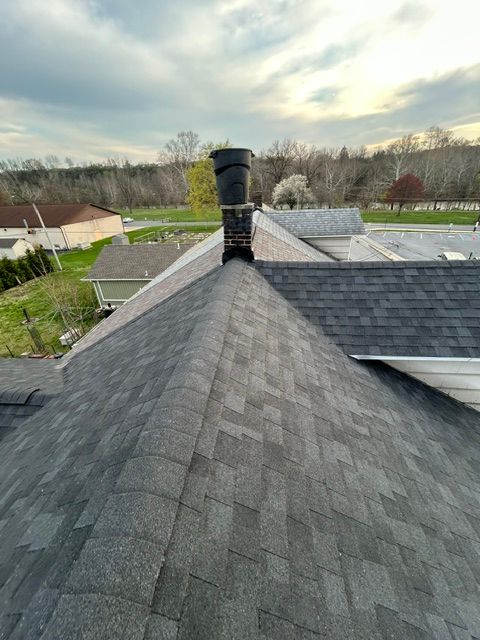 View from a shingled rooftop toward a chimney, neighboring houses, and a cloudy sunset sky