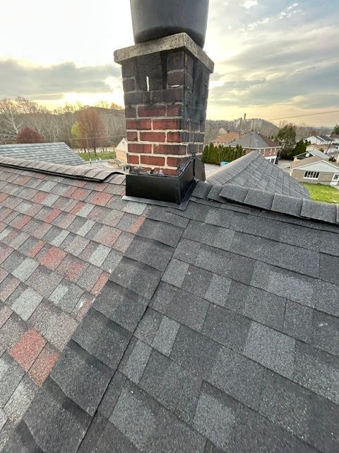 Rooftop with a brick chimney and dark shingled roof at sunset, overlooking nearby houses and trees
