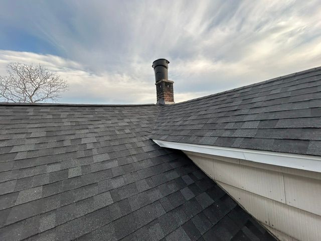 Rooftop with dark shingles, white gutter, and a brick chimney under a cloudy sky