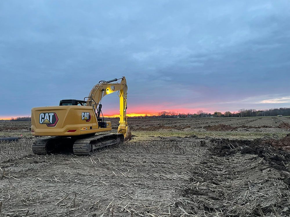 A cat excavator is sitting in a field at sunset.